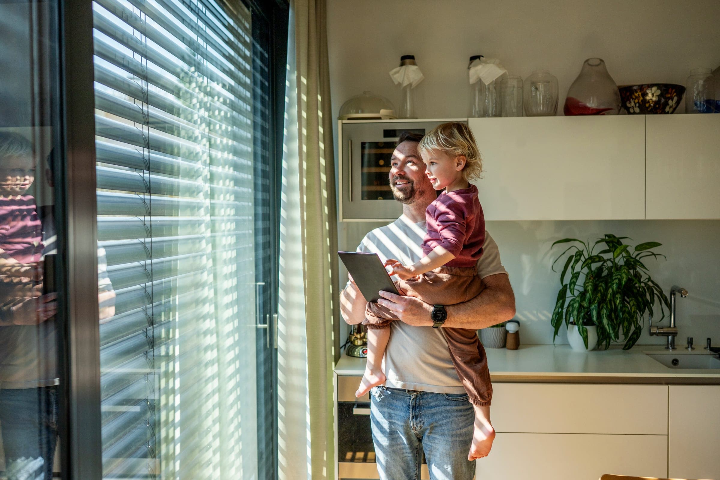 Vater mit Kind am Fenster steuert die Raffstore-Lamellen im Streiflicht per Tablet